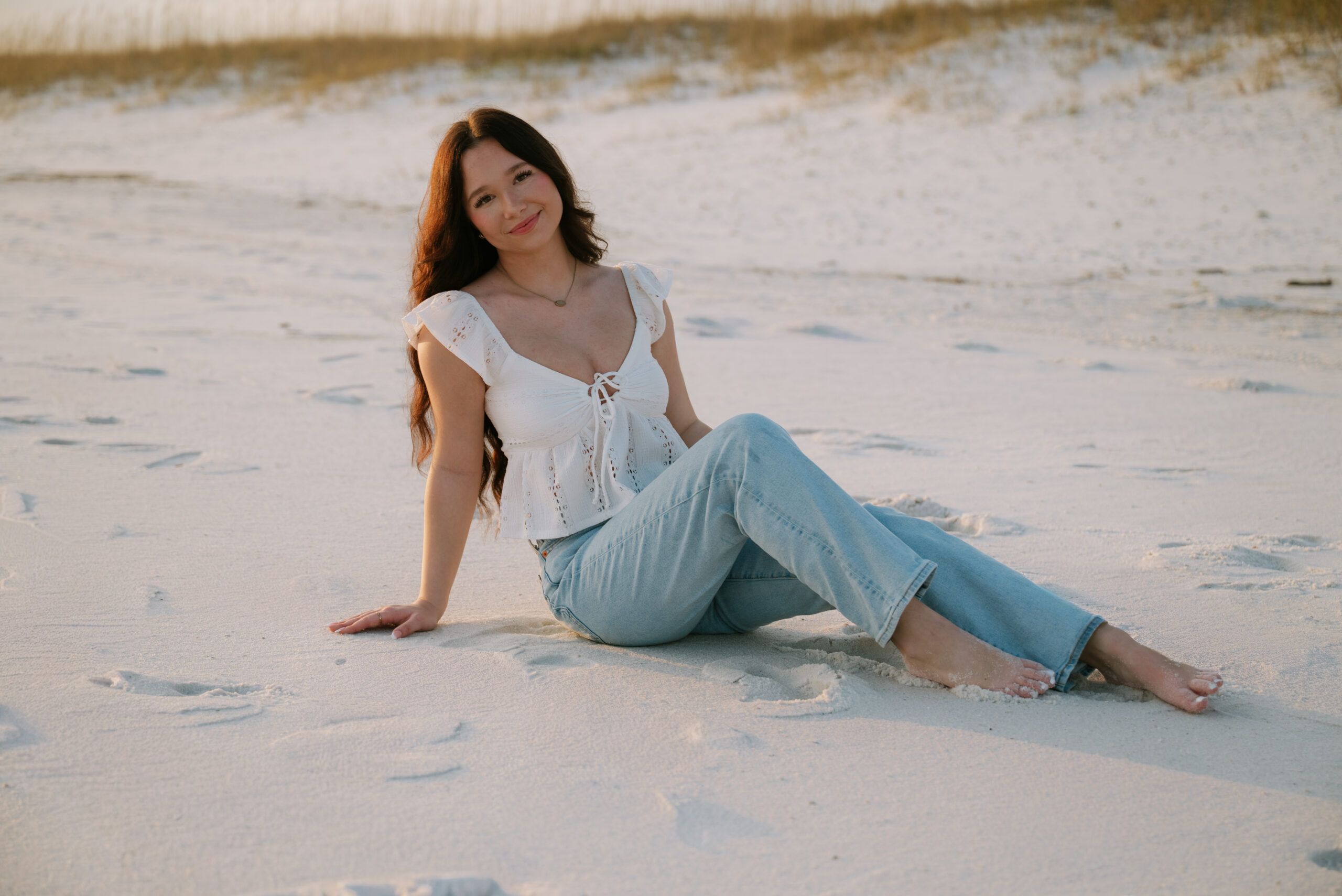 pensacola senior photographer | a girl in jeans and a white top sitting down in the sand on Pensacola Beach
