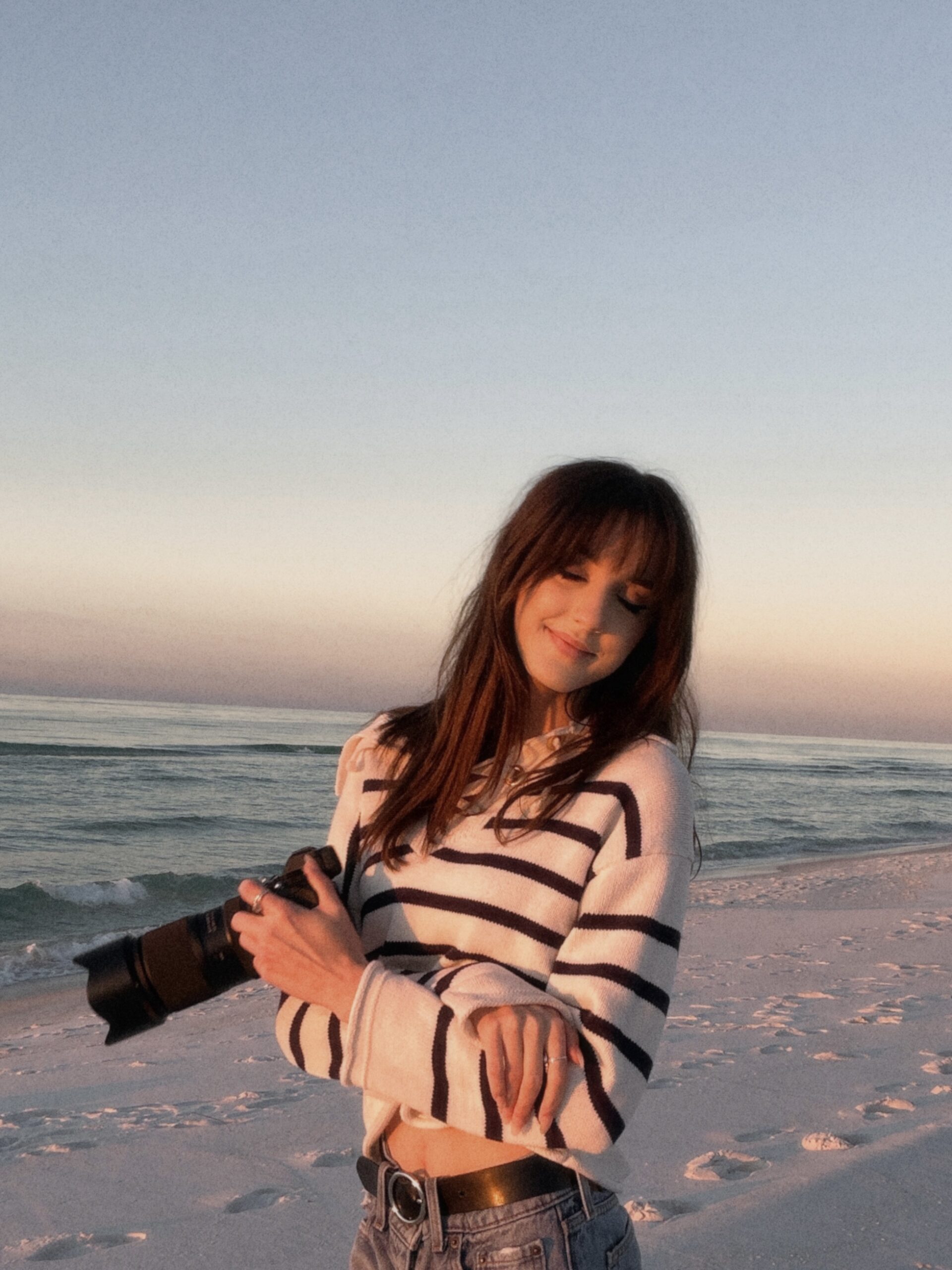 Rowan is on Pensacola Beach. Her camera is in one of her hands and her arms are crosses as she rests her chin on her shoulder and smiles with her eyes closed. The ocean is behind her and the sun is setting. Pensacola Beach.