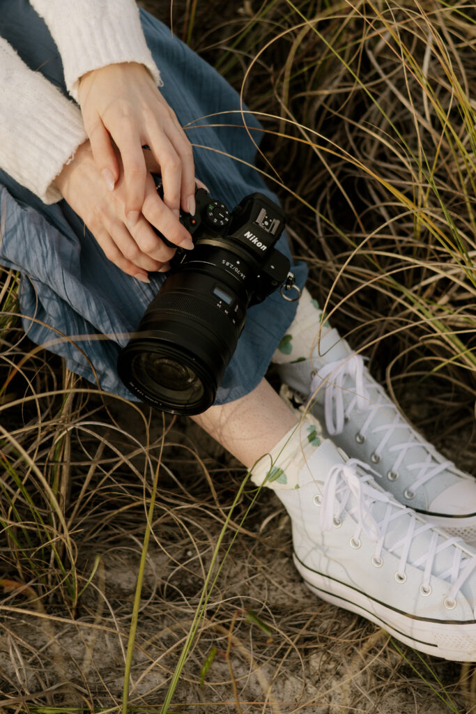 Pensacola couples photographer sitting in a field at Fort Pickens on the Florida Panhandle. A detail closeup of her holding her camera by her legs.