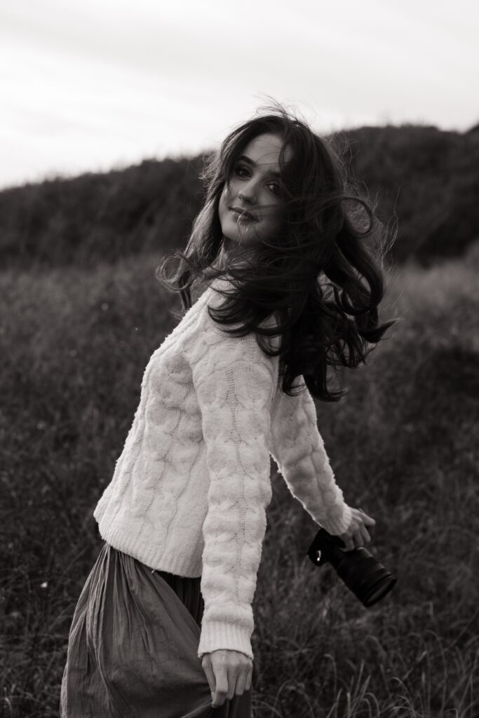 Pensacola couples photographer spinning in a field at Fort Pickens on the Florida Panhandle. Her hair is blowing as she holds her camera with her arms out. Black and white.