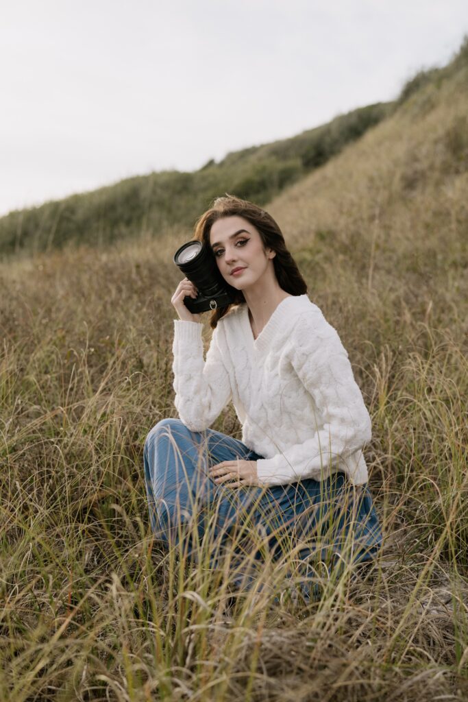 Pensacola couples photographer. Sitting in a field of grass at Fort Pickens wearing a cream sweater and holding her camera up by her face.