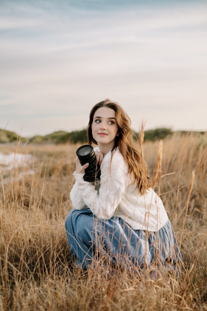 Pensacola couples photographer crouching in a field at Fort Pickens on the Florida Panhandle. She is looking over her shoulder as she holds her camera and smiles.
